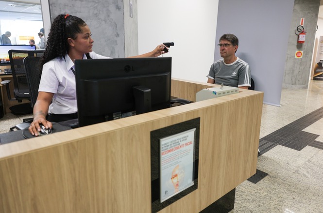 A imagem mostra uma cena em um ambiente interno, possivelmente um escritório ou recepção. Há uma pessoa sentada atrás de um balcão de madeira com um computador, e ela está segurando um dispositivo, talvez uma câmera ou scanner, apontado para uma pessoa que está sentada do outro lado do balcão. A segunda pessoa está em uma cadeira, usando uma camiseta cinza. No balcão, há um cartaz sobre reconhecimento facial. O ambiente parece moderno, com piso claro e sinalização na pared