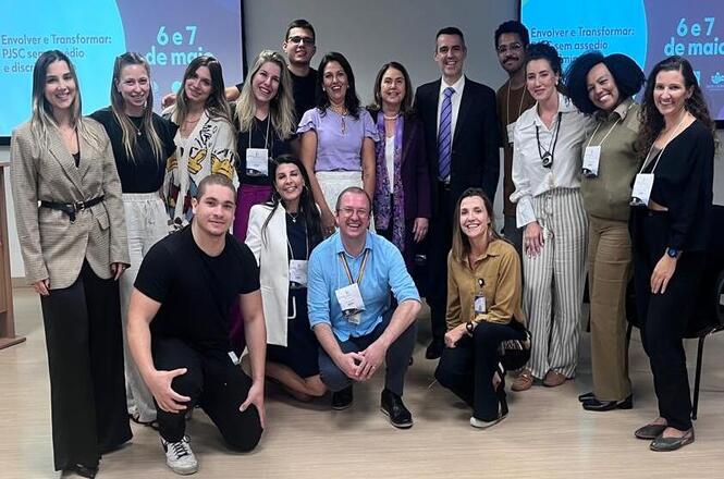 A imagem mostra um grupo de pessoas posando para uma foto em um ambiente que parece ser uma sala de conferências ou reunião. Doze pessoas estão em pé, enquanto três estão agachadas ou ajoelhadas na frente. Ao fundo, há uma tela de projeção com texto parcialmente visível, mencionando "6 e 7 de maio" e o tema "assédio e discriminação". As pessoas estão usando crachás, sugerindo que estão participando de um evento ou conferência. Todos parecem felizes ou sorridentes na foto.