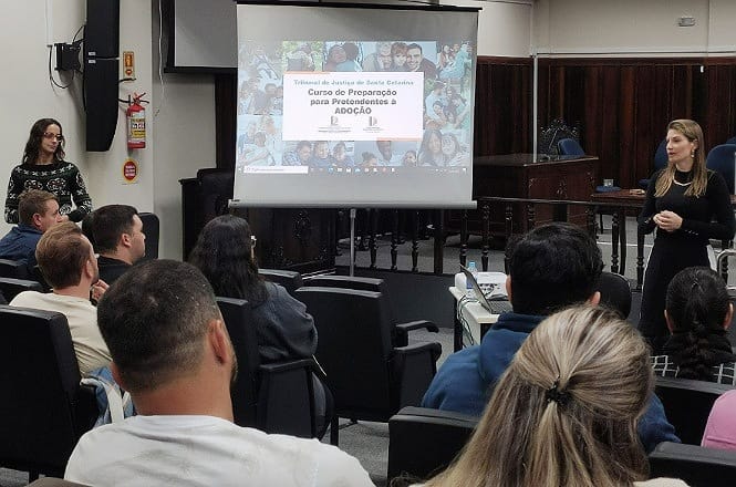 A imagem mostra uma sala de aula ou auditório. Há uma apresentação sendo projetada em uma tela com o título "Curso de Preparação para Pretendentes à Adoção" pelo Tribunal de Justiça de Santa Catarina. Algumas pessoas estão sentadas assistindo à apresentação, enquanto duas apresentadoras estão em pé à frente. Uma delas está falando ao público. O ambiente tem cadeiras organizadas em fileiras e detalhes de madeira nas paredes.