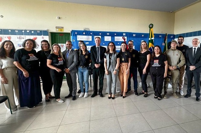 A imagem mostra um grupo de pessoas posando para uma foto em um ambiente interno. Há várias pessoas de pé, alinhadas em um semicírculo. Algumas estão vestindo roupas formais, enquanto outras usam uniformes ou camisetas temáticas. Ao fundo, há cartazes com logotipos e um quadro azul. Também é possível ver a bandeira do Brasil. O ambiente parece ser de um evento ou cerimônia oficial.