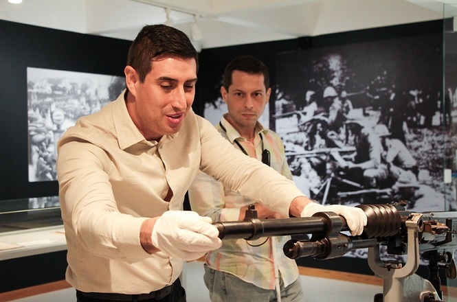 Na imagem, há duas pessoas. A pessoa à esquerda segura uma arma antiga montada em um suporte e está usando luvas brancas. Ao fundo, há uma grande foto em preto e branco na parede que parece mostrar uma cena histórica de soldados em combate. Ambos estão em um ambiente que parece ser uma exposição ou museu.