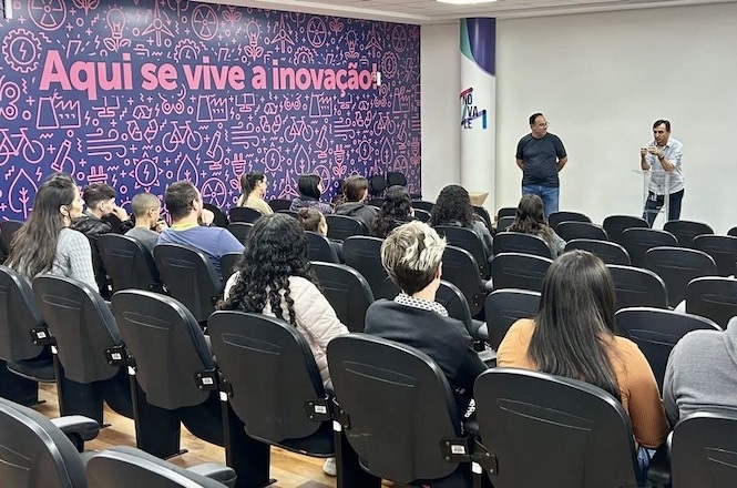 A imagem mostra uma sala de conferências com várias pessoas sentadas em cadeiras, assistindo a uma apresentação de dois palestrantes que estão em pé na frente da sala. A parede ao fundo apresenta um mural roxo com desenhos e a frase "Aqui se vive a inovação!" em destaque. A sala tem um ambiente moderno e parece estar relacionada a temas de tecnologia ou inovação.