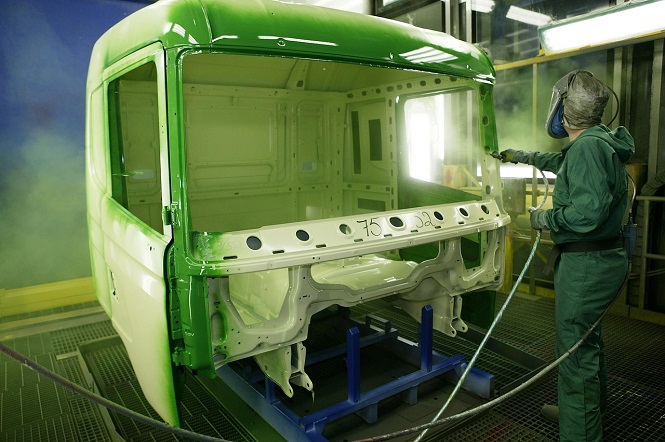 A imagem mostra uma cabine de veículo sendo pintada. Um pintor está aplicando tinta verde na estrutura usando uma pistola de pulverização. A cabine está sobre um suporte e o ambiente parece ser uma área industrial ou uma oficina de pintura, com iluminação adequada e equipamentos de proteção usados pelo trabalhador.