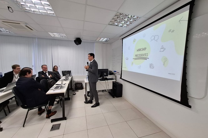 A imagem mostra uma sala de conferência ou aula. Um homem está falando em pé, segurando um microfone, ao lado de uma tela de projeção na qual está exibida a apresentação intitulada “WORKSHOP PRECEDENTES”. Há quatro pessoas sentadas em frente a ele, aparentemente ouvindo a apresentação. A sala tem um ambiente corporativo com paredes brancas e teto com luzes embutidas.