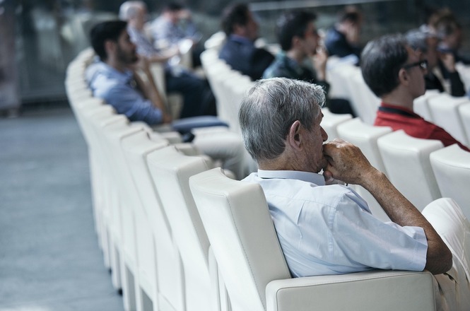 A imagem mostra várias pessoas sentadas em cadeiras de um auditório ou sala de conferências. Eles parecem estar assistindo a uma apresentação ou palestra. As cadeiras são brancas, e o ambiente é bem iluminado. As pessoas estão vestidas de maneira casual ou semi-formal. A cena transmite uma atmosfera de concentração e atenção.