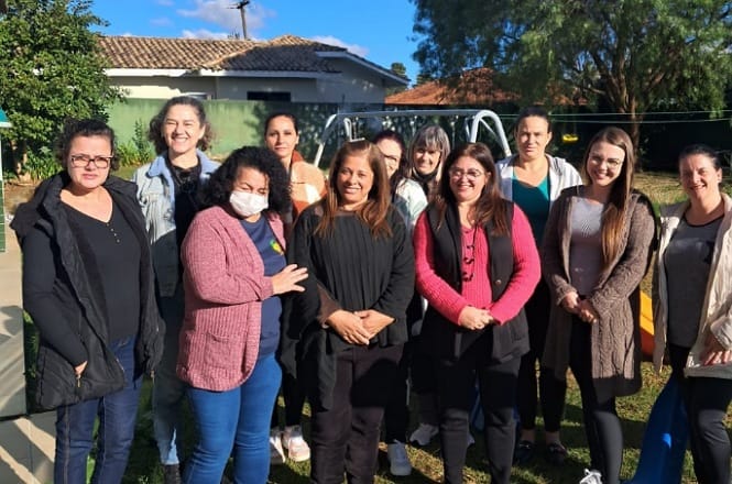 A imagem mostra um grupo de doze pessoas, principalmente mulheres, reunidas ao ar livre. Algumas estão sorrindo, e uma delas está usando máscara. Ao fundo, há uma casa com telhado de telhas, árvores e um balanço. O céu está claro e há uma sensação de convivência alegre entre as pessoas.