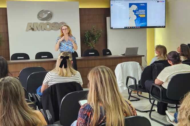 A imagem mostra uma mulher fazendo uma apresentação em uma sala de conferências. Ela está de pé, falando para uma plateia de pessoas sentadas em cadeiras. Atrás dela, há uma mesa com cadeiras e um letreiro na parede com o texto "AMOSC". Ao lado, há uma tela exibindo uma apresentação eletrônica. As pessoas na audiência parecem estar prestando atenção na apresentação. A sala possui uma decoração simples e moderna, com plantas ao fundo.