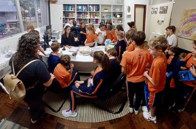 Na imagem, vemos um grupo de crianças vestindo uniformes escolares laranja e azul. Elas estão reunidas em torno de uma mesa em um ambiente que parece ser um escritório ou sala de estudos. Duas mulheres estão presentes, provavelmente supervisionando ou ensinando as crianças. Ao fundo, há uma estante cheia de livros e alguns objetos decorativos. As crianças parecem estar atentas e interessadas no que está sendo explicado. O local tem janelas grandes, permitindo a entrada de luz natural.