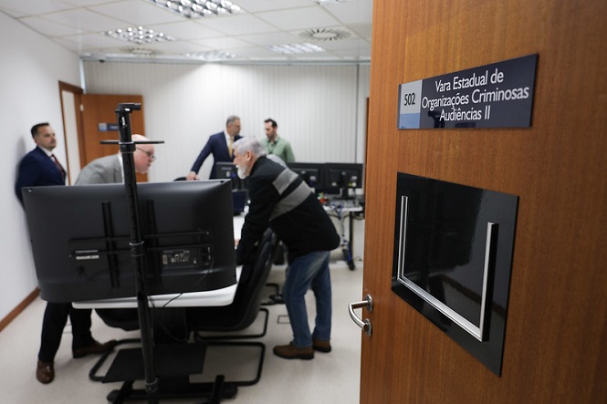 A imagem mostra uma sala com várias pessoas dentro, discutindo ou trabalhando em frente a computadores. A porta da sala está aberta e há uma placa ao lado que diz "Vara Estadual de Organizações Criminosas Audiências II". A sala parece ser parte de uma instalação oficial ou governamental, possivelmente ligada ao sistema judiciário. Há várias cadeiras e monitores no ambiente.