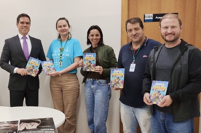 A imagem mostra cinco pessoas posando para a câmera, cada uma segurando um exemplar de um livro ou revista com uma capa colorida. Quatro homens e uma mulher estão de pé em frente a uma mesa branca. A mulher no centro veste uma camiseta azul clara e calça bege, e as outras pessoas estão vestidas de maneira casual ou formal. Ao fundo, há uma parede clara com uma porta de madeira e uma placa. A imagem parece ter sido tirada em um ambiente interno, possivelmente em um escritório ou local de reunião.
