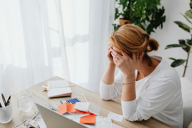 A imagem mostra uma mulher sentada à mesa com a cabeça entre as mãos, parecendo preocupada ou estressada. Na mesa, há um laptop, papéis, notas adesivas, um bloco de anotações, dinheiro e um copo d'água. Ao fundo, há uma cortina clara e algumas plantas. A cena sugere um ambiente de trabalho ou escritório em casa.