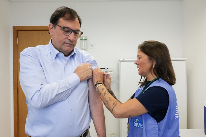 Na imagem, há um homem de óculos e camisa azul sendo vacinado por uma profissional de saúde. Ele está levantando a manga da camisa, enquanto a profissional, em um colete azul com a palavra "VIGILÂNCIA", administra a vacina. O cenário parece ser uma sala de vacinação, com uma porta de madeira e um armário branco ao fundo.