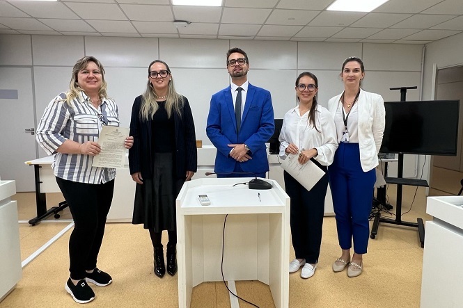 Na imagem, há cinco pessoas posando para a foto em uma sala que parece ser um ambiente de conferência ou reunião. Três mulheres estão vestidas de forma formal com roupas como blazers e camisas, e um homem veste um terno azul. Há também uma mulher segurando um papel. No ambiente, é possível ver um púlpito branco na frente, além de uma televisão ou monitor ao fundo. A sala tem iluminação branca e um teto com painéis.