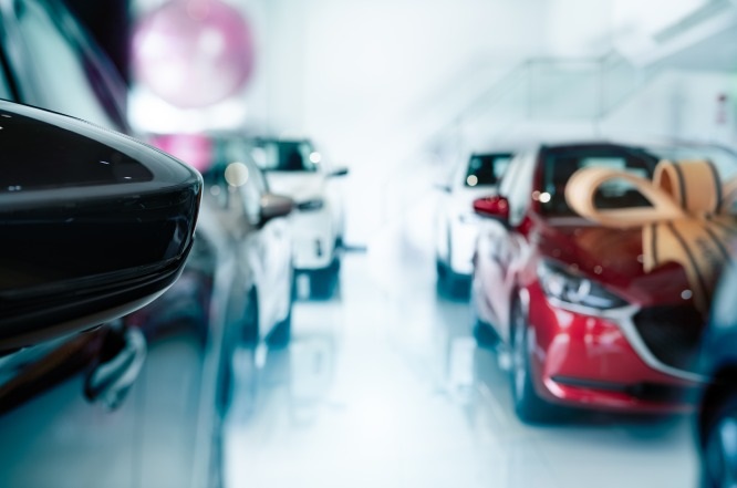 A imagem mostra o interior de uma concessionária de automóveis, onde diversos carros estão estacionados. Em primeiro plano, há uma parte de um espelho lateral de um carro. Ao fundo, é possível ver outros veículos, incluindo um carro vermelho à direita com um grande laço sobre ele, sugerindo que pode ser um presente ou uma promoção especial. O ambiente é bem iluminado e moderno, com reflexos brilhando no chão.