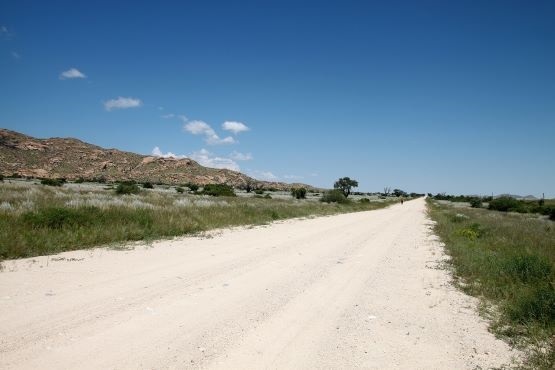 A imagem mostra uma estrada de terra, reta, sob um céu claro e azul. Há vegetação rasteira ao longo da estrada, com algumas árvores espalhadas. Ao fundo, pode-se ver uma cadeia de montanhas de tom marrom-claro. O cenário sugere uma área rural ou de savana.