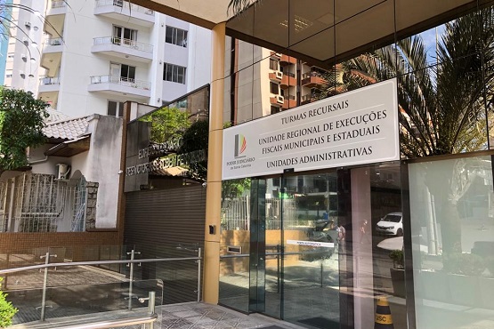 A imagem mostra a entrada de um prédio com uma fachada de vidro. Na fachada, há uma placa com a inscrição "TURMAS RECURSAIS, UNIDADE REGIONAL DE EXECUÇÕES FISCAIS MUNICIPAIS E ESTADUAIS, UNIDADES ADMINISTRATIVAS". Ao lado da porta, há a indicação de um acesso para pessoas com deficiência. Ao fundo, é possível ver parte de outro edifício e algumas plantas próximas à entrada.