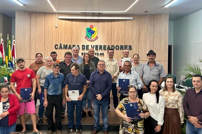 A imagem mostra um grupo de pessoas posando para uma foto em um ambiente formal. Elas estão em uma sala com um púlpito ao fundo, onde se lê "Câmara de Vereadores Benedito Novo - SC". Algumas pessoas seguram pastas ou documentos e há tanto homens quanto mulheres no grupo. No canto da imagem, podem ser vistas bandeiras e plantas ornamentais. O local parece ser um espaço destinado a eventos ou reuniões oficiais.