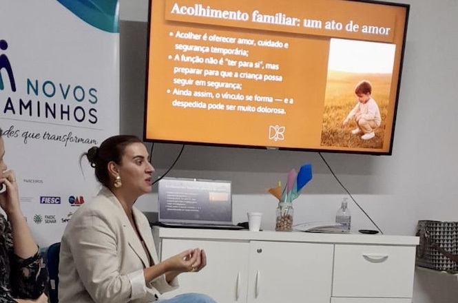 A imagem mostra uma mulher sentada em uma sala enquanto realiza uma apresentação sobre "Acolhimento familiar: um ato de amor". Atrás dela, há uma tela exibindo um slide com tópicos sobre acolhimento e segurança temporária para crianças, enfatizando a importância de oferecer amor e cuidado. Ao lado da tela, há um móvel com objetos como copos e materiais coloridos. O ambiente parece ser uma sala de conferências ou de aula.