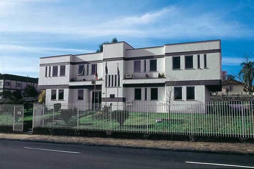 A imagem mostra um edifício de dois andares com um estilo arquitetônico moderno. A construção é predominantemente branca, com linhas retas e janelas retangulares. Há um pequeno jardim na frente, cercado por uma cerca metálica. Na frente do edifício, há uma calçada e a rua. O céu está azul, sugerindo um dia claro e ensolarado.