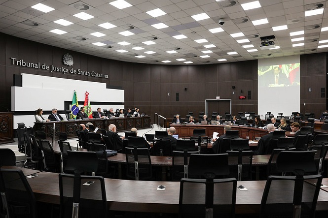 A imagem mostra uma sala de tribunal do Tribunal de Justiça de Santa Catarina. Há várias pessoas sentadas em uma disposição circular ao longo de uma mesa grande, que parece ser o plenário. Na parte superior esquerda, está escrito "Tribunal de Justiça de Santa Catarina" e há bandeiras do Brasil e provavelmente de Santa Catarina. As pessoas parecem estar participando de uma sessão ou reunião formal. O ambiente é bem iluminado com várias luzes no teto, e há uma projeção na parede à direita.