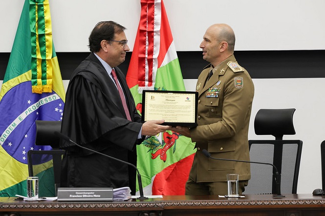 Na imagem, há duas pessoas em pé, uma entregando um certificado para a outra. À esquerda, está um homem vestindo uma toga preta e uma gravata vermelha, enquanto à direita, há um homem em uniforme militar marrom. Eles estão em um ambiente que parece ser uma sala oficial ou cerimonial. Atrás deles, há bandeiras do Brasil e de um estado brasileiro. Uma placa com nome está visível na mesa em frente à pessoa à esquerda.