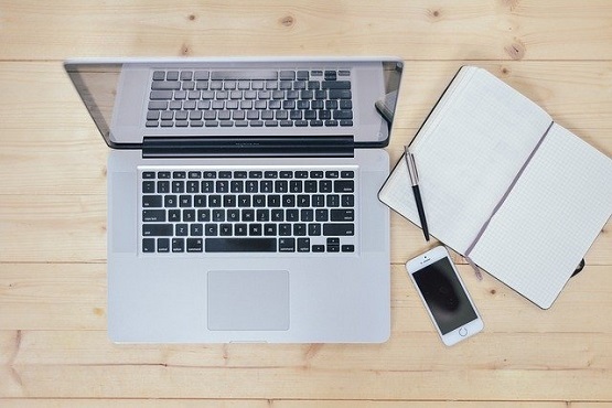A imagem mostra um espaço de trabalho com um laptop prateado aberto sobre uma mesa de madeira clara. Ao lado do laptop, há um caderno aberto com capa preta e folhas quadriculadas, acompanhado de uma caneta preta. Também há um smartphone branco, virado com a tela para cima, posicionado ao lado do caderno.