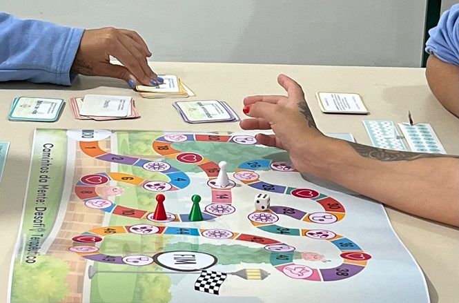 Na imagem, duas pessoas estão jogando um jogo de tabuleiro. O tabuleiro é colorido com caminhos e desenhos de corações e cérebros. Há peões de cores diferentes no tabuleiro e cartas sendo manuseadas pelos jogadores. O tabuleiro tem uma linha de chegada com bandeira de chegada preta e branca. Parece ser um jogo de desafios relacionados à mente e ao bem-estar.