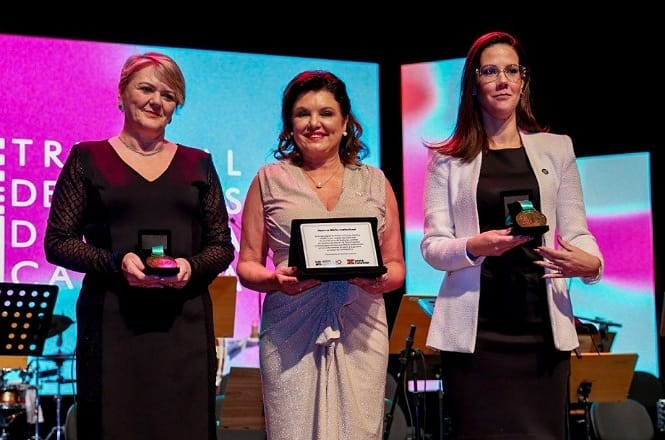 Na imagem, há três mulheres em pé em um palco, todas segurando prêmios. A mulher à esquerda está usando um vestido preto, a do meio está usando um vestido claro e segurando uma placa, e a mulher à direita está vestindo um blazer branco com um vestido preto por baixo. Ao fundo, há uma tela com projeções coloridas e instrumentos musicais, sugerindo um evento ou cerimônia de premiação.