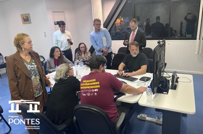 A imagem mostra um grupo de pessoas em uma sala de reuniões. Elas estão reunidas em torno de uma mesa que possui computadores e monitores. Uma pessoa está sentada manipulando um dos computadores, enquanto outras pessoas obseravam de pé e sentadas ao redor. A atmosfera parece ser de concentração, possivelmente discutindo ou analisando algo no computador. As pessoas estão vestidas de maneira variada, algumas em trajes mais casuais e outras em trajes formais. Há uma janela grande ao fundo e algumas cadeiras adicionais na sala.