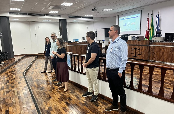 Na imagem, há seis pessoas em pé, aparentemente participando de um evento ou apresentação. Eles estão em um ambiente interno, que parece ser uma sala de conferências ou auditório, com piso de madeira e um projetor exibindo algo em uma tela ao fundo. Alguns deles seguram microfones, indicando que estão falando ou se apresentando para uma audiência. Há também bandeiras ao fundo, que podem representar instituições ou localidades. O local parece ser formal, sugerindo um evento profissional ou educacional.