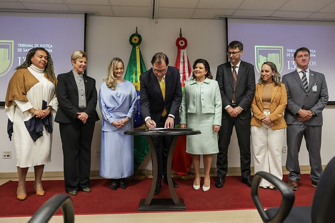 Na imagem, há um grupo de oito pessoas em pé, em frente a uma parede com duas bandeiras, possivelmente do Brasil e de Santa Catarina. Eles estão em um ambiente formal, provavelmente em uma cerimônia ou evento oficial. No centro, uma pessoa está assinando um documento sobre uma mesa. As pessoas ao redor estão vestidas formalmente. Ao fundo, há telas eletrônicas com o texto "Tribunal de Justiça de Santa Catarina" indicando uma ocasião institucional.