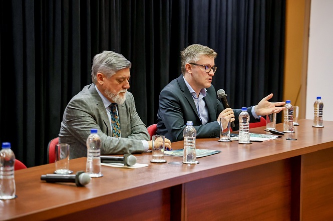 A imagem mostra duas pessoas sentadas em uma mesa de conferência. Um deles segura um microfone e parece estar falando, enquanto o outro está olhando para baixo, talvez lendo ou fazendo anotações. Na mesa, há várias garrafas de água e microfones. O fundo é composto por uma cortina preta. Ambos os homens estão vestindo ternos.