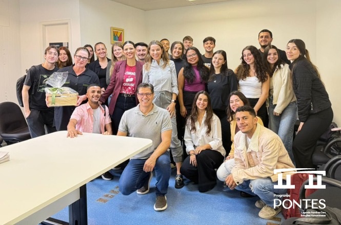 A imagem mostra um grupo de pessoas reunidas em uma sala. Elas estão sorrindo e posando para a foto. Há uma mesa branca à esquerda e, ao fundo, uma parede branca com uma pequena obra de arte pendurada. As pessoas parecem estar em um ambiente de trabalho ou educativo. Há também um logotipo no canto inferior direito da imagem com o texto "Programa Pontes".