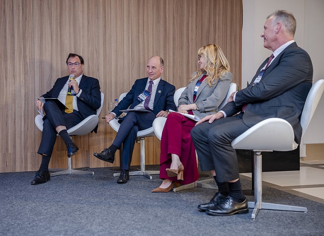 A imagem mostra quatro pessoas sentadas em cadeiras de um auditório, participando de um painel de discussão ou conferência. Eles estão em um ambiente corporativo ou de negócios. Uma pessoa à esquerda está segurando um microfone, provavelmente como moderador ou apresentador. As pessoas estão vestidas de forma formal, com ternos e roupas apropriadas para o evento. A parede de fundo é revestida com madeira, proporcionando uma aparência sofisticada ao ambiente.