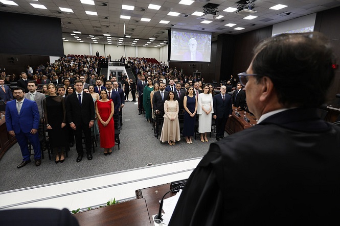 A imagem mostra uma cerimônia em um auditório grande e bem iluminado. No primeiro plano, um homem em trajes formais (aparentemente uma toga) está em pé, observando a plateia. À frente dele, várias pessoas estão alinhadas em pé, vestidas formalmente, com vestidos e ternos. A plateia, ao fundo, também está cheia, indicando que o evento é formal e possui muitos participantes. No fundo, há um telão exibindo uma imagem. Todo o ambiente tem uma aparência profissional e solene.