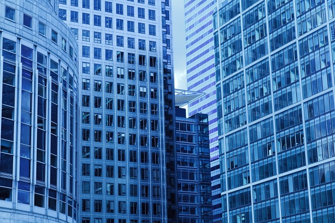 A imagem mostra vários arranha-céus modernos de vidro e metal cercados por outras construções urbanas. Os prédios exibem principalmente tons de azul e apresentam diversas janelas, refletindo a luz e o céu. As estruturas têm um design contemporâneo, típico de um centro de negócios urbano.