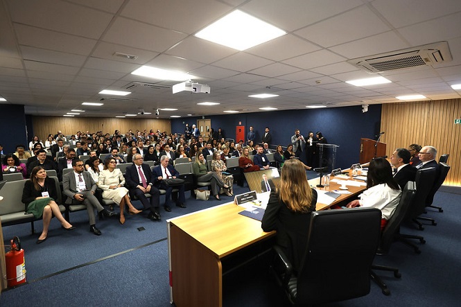 A imagem mostra uma sala de conferências onde muitas pessoas estão sentadas assistindo a uma apresentação ou palestra. Na frente, há uma mesa com várias pessoas sentadas, provavelmente palestrantes ou membros de um painel. O público parece estar atento, e o ambiente é bem iluminado e climatizado, com teto de gesso e luzes embutidas. Há também equipamentos de projeção visíveis no teto. A sala tem cadeiras organizadas em fileiras, criando um ambiente formal e profissional.