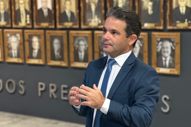 A imagem mostra um homem de terno escuro e gravata clara falando em um ambiente interior. Ao fundo, há uma parede com fotos emolduradas de pessoas em formato de galeria. O fundo também possui letras grandes parcialmente visíveis na parede. O homem está gesticulando com as mãos enquanto parece estar em algum tipo de apresentação ou discurso.