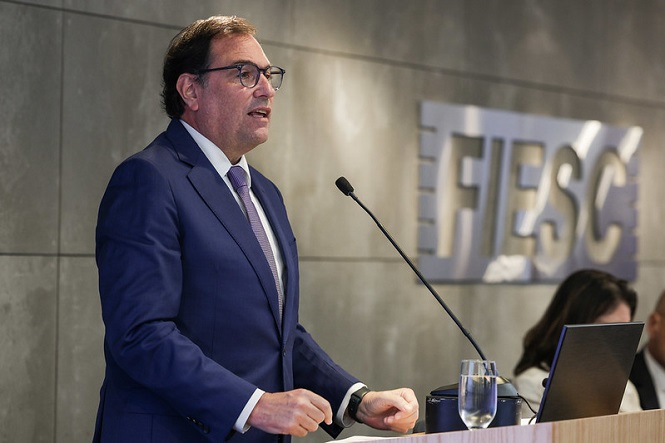 A imagem mostra um homem de terno azul, gravata roxa e óculos, falando ao microfone em um púlpito. Ele está em um ambiente profissional, provavelmente durante uma apresentação ou discurso. Ao fundo, vê-se uma parede com a inscrição "FIESC". Há também um copo de água sobre o púlpito e outra pessoa, desfocada, ao fundo.