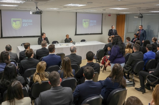 A imagem mostra o interior de uma sala de tribunal. Várias pessoas estão posicionadas à frente, algumas de pé, atrás de uma bancada elevada. Entre elas, estão funcionários de segurança. Algumas pessoas estão sentadas na plateia, de costas para a câmera. Na parede ao fundo, há um crucifixo. O ambiente parece ser formal e institucional.