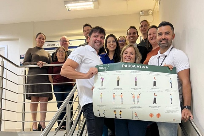 A imagem mostra um grupo de pessoas posando em uma escada. Eles seguram um cartaz intitulado "PAUSA ATIVA", que parece apresentar exercícios ou instruções de alongamento. O grupo está sorrindo, e a cena ocorre em um ambiente interno, possivelmente em uma instituição ou escritório. Ao fundo, há uma placa que diz "Fórum".