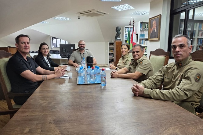 A imagem mostra seis pessoas sentadas ao redor de uma mesa de madeira em uma sala de conferências ou reunião. Na mesa, há várias garrafas de água e copos. As pessoas estão usando roupas formais; algumas estão com uniformes militares. Ao fundo, há prateleiras com livros, uma bandeira, um busto, e um retrato pendurado na parede. A sala parece ser bem iluminada, com teto inclinado e luz natural entrando pelas janelas.