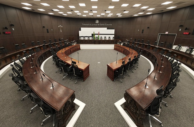 A imagem mostra uma sala de tribunal moderna e ampla, com um design interior sofisticado. As mesas são dispostas em um formato de semicírculo, equipadas com microfones para uso dos bancadas. As cadeiras pretas estão alinhadas ao redor das mesas. Na parede ao fundo, há um painel que indica que este é o Tribunal de Justiça de Santa Catarina. O ambiente é bem iluminado, com um teto cheio de luminárias embutidas. Também é notável a presença de um crucifixo na parede. A bandeira brasileira é visível em um canto elevado ao fundo da sala.
