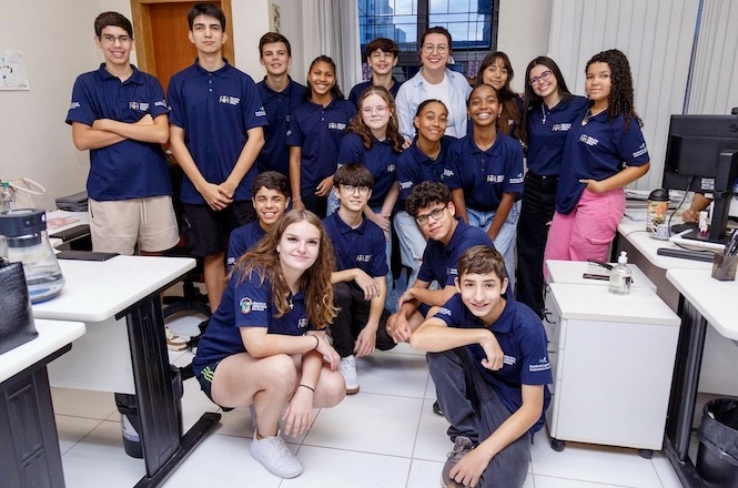 A imagem mostra um grupo de jovens em uma sala. Eles estão vestidos com camisetas azul marinho, algumas com logotipos. A maioria está de pé, enquanto alguns estão agachados na frente. A sala parece ser um ambiente de escritório, com mesas, cadeiras e computadores. Todos estão sorrindo e parecem estar participando de algum tipo de evento ou reunião em grupo.