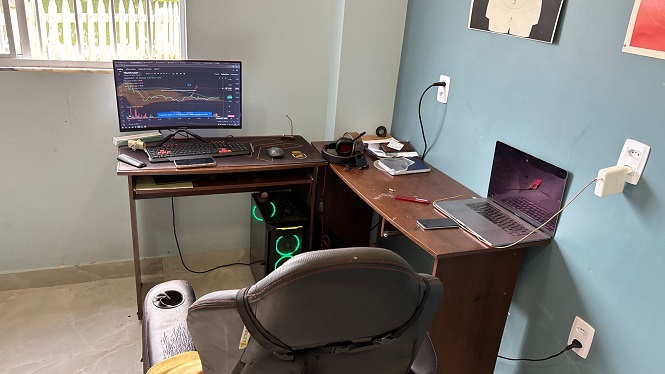 A imagem mostra um ambiente de escritório com duas mesas em L encostadas na parede azul. Na mesa à esquerda, há um monitor com um gráfico exibido, um teclado, um mouse e uma CPU abaixo da mesa. Na mesa à direita, há um laptop aberto, um caderno, um celular e alguns cabos conectados na tomada próxima. Também há uma cadeira preta em frente às mesas e uma janela à esquerda que permite a entrada de luz natural. Há um quadro pendurado na parede acima das mesas.