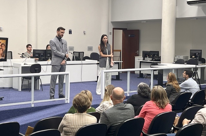 A imagem mostra duas pessoas apresentando em uma sala, possivelmente uma sala de conferências ou tribunal, diante de um grupo de pessoas sentadas. A pessoa à esquerda está usando um terno cinza, e a pessoa à direita está vestindo roupa branca e cinza. Atrás deles, há mais pessoas sentadas em uma bancada com computadores, e há uma porta ao fundo. O ambiente é profissional, com pessoas prestando atenção à apresentação.