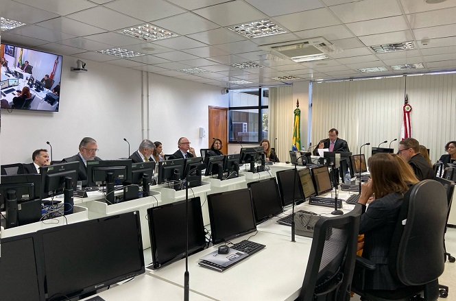 A imagem mostra uma sala de reuniões com várias pessoas sentadas ao redor de uma mesa em formato de U. Há vários computadores e monitores presentes na mesa. As pessoas parecem estar participando de uma reunião ou discussão. Na parede, há uma tela exibindo o que parece ser uma videoconferência. Além disso, são visíveis bandeiras na sala, indicando talvez um ambiente oficial ou governamental. A sala é bem iluminada, com um teto de iluminação embutida.