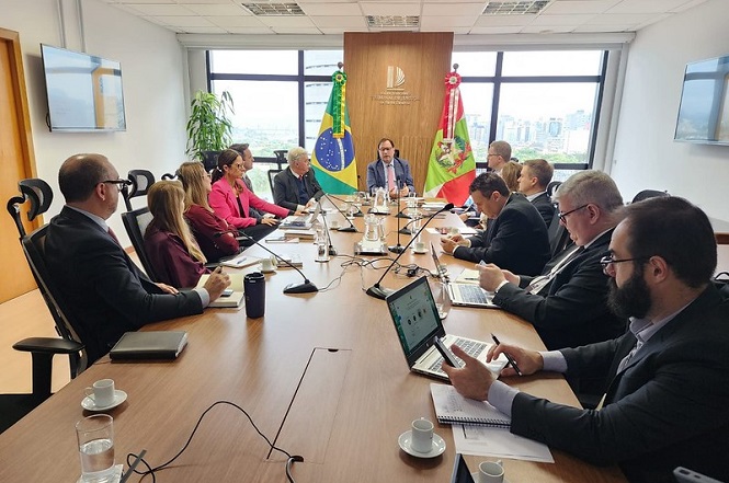 A imagem mostra uma reunião em uma sala de conferências. Há várias pessoas sentadas ao redor de uma mesa comprida, algumas usando ternos. No centro da mesa há microfones e papéis. Duas bandeiras estão na parede ao fundo, uma do Brasil e outra à direita, com um brasão. O ambiente parece ser formal e decorado com algumas plantas. Existem laptops e cadernos em uso na mesa, e há copos de café e água sobre ela. A sala tem janelas grandes que mostram a paisagem urbana.