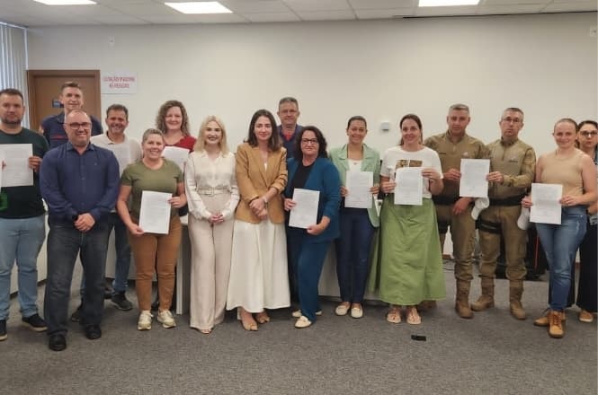 A imagem mostra um grupo de pessoas em pé, lado a lado, posando para uma foto em uma sala. Algumas pessoas estão segurando documentos nas mãos. Todos parecem estar sorrindo. A sala tem iluminação de teto e uma porta ao fundo. Há uma variação de vestimentas entre os presentes, incluindo roupas formais e uniformes. A atmosfera parece ser de um evento ou reunião formal.