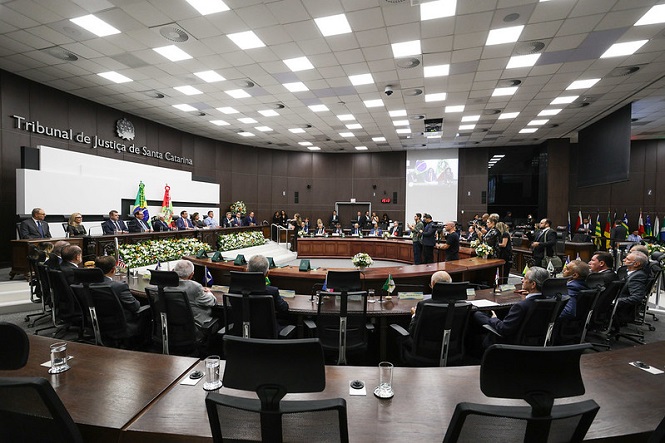 A imagem mostra uma grande sala de conferências ou um tribunal com diversas pessoas sentadas ao redor de uma mesa em formato oval. No fundo, está escrito "Tribunal de Justiça de Santa Catarina". Há bandeiras de diversos estados nas laterais. As pessoas presentes estão em trajes formais e parecem estar participando de um evento oficial ou sessão formal. Há arranjos de flores na mesa, e algumas pessoas estão em pé, possivelmente fazendo um discurso ou apresentação.